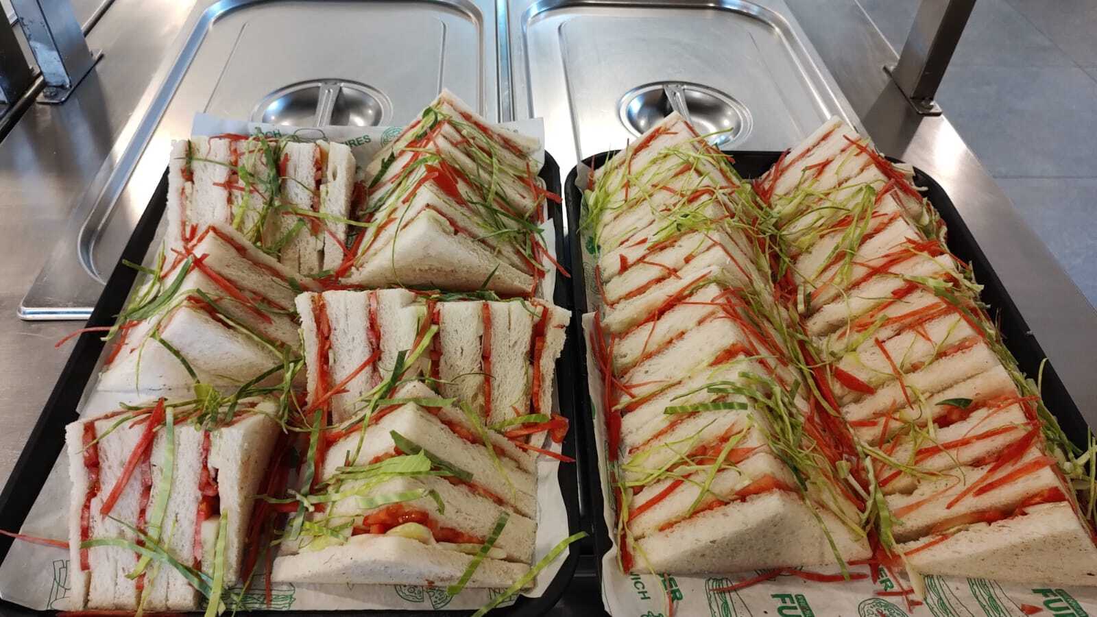 Two trays of triangle-cut sandwiches with tomato and shredded carrot and cabbage, on a steel service counter.