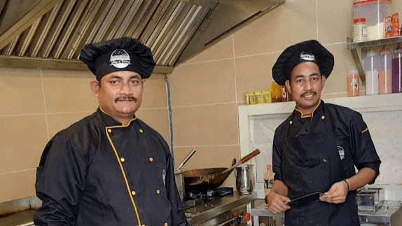 Two chefs in black kitchen whites and branded caps standing beside the range in the kitchen.